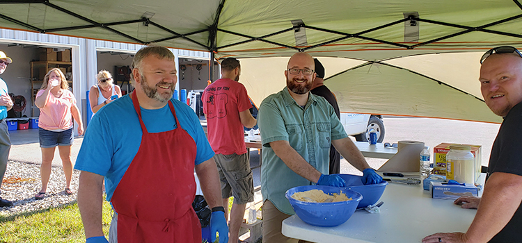 A few men smile at the camera while preparing food in large bowls, underneath a large event canopy.