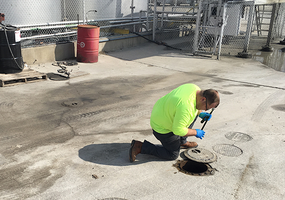 A man kneels on pavement and looks into an open maintenance access pipe with the manhole slid to one side.