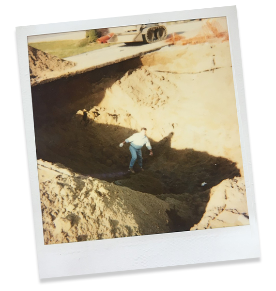 Vintage polaroid of a man standing in a deep hole of soil, with a small front loader in the background.