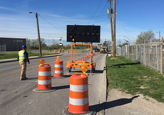 A man in construction gear walks on a road alongside a line of construction barrels, traffic cones, and an LED arrow sign.